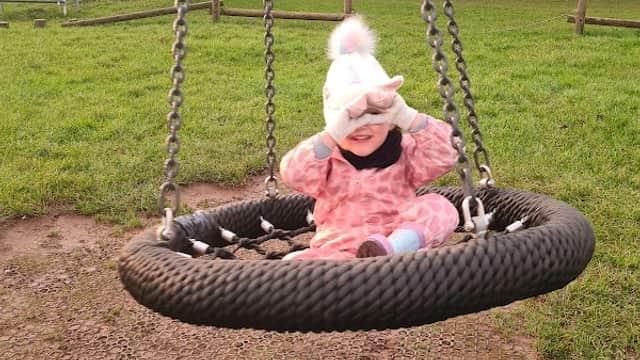 A child in a pink outfit sits on a large swing, smiling and shielding their eyes from the sun in a grassy park.