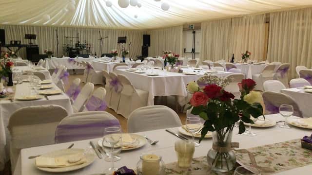 Elegant dining setup with tables adorned in white linens, purple accents, floral centerpieces, and soft lighting.