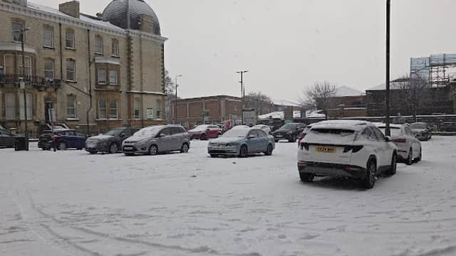 Snow-covered car park with parked vehicles and a historic building in the background under a gray sky.