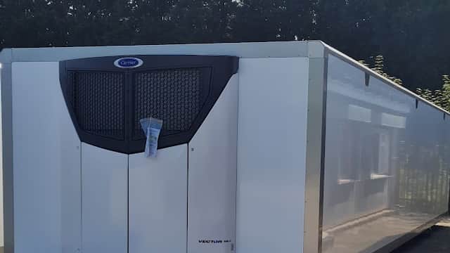 White refrigerated container with a black ventilation grille, parked in a sunny area surrounded by trees.