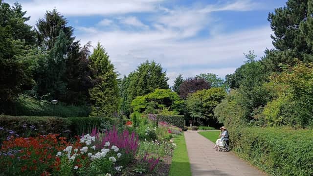 Lush greenery and colorful flower beds line a pathway; a person sits on a bench under a blue sky with fluffy clouds.
