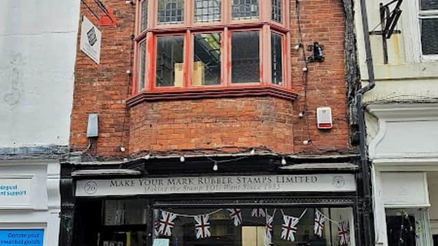Historic brick shopfront with large windows, featuring "Make Your Mark RUBBER STAMP" sign and colorful display.