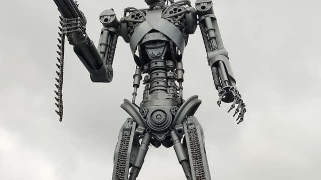 A large metallic robot statue holding a gun, standing on a platform against a cloudy sky.