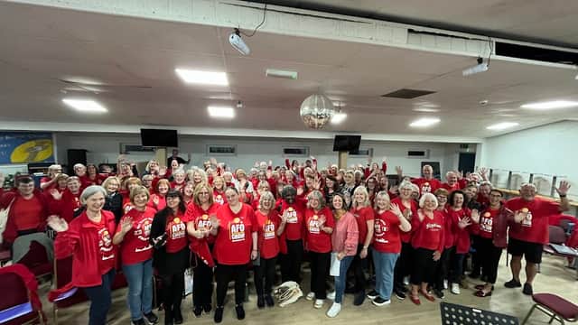 A large group of people in red shirts, smiling and waving, gathered in a spacious hall with chairs and a disco ball.