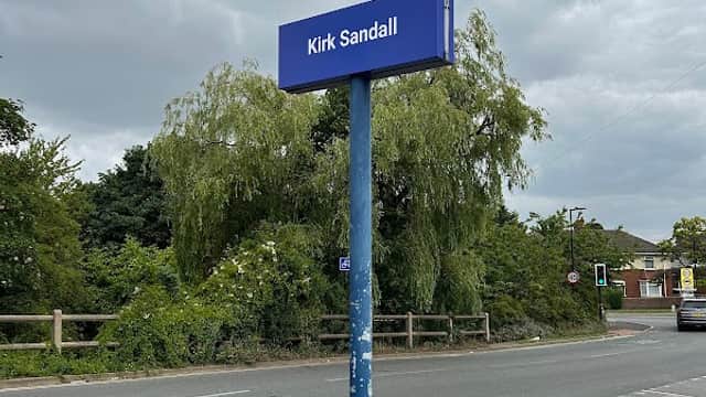 Kirk Sandall station sign on a pole near a car park, surrounded by greenery and cloudy skies.