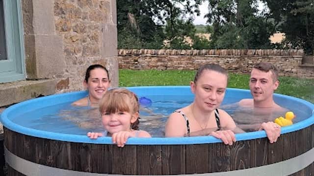 Four people relax in a wooden hot tub outside a stone farmhouse, surrounded by greenery.