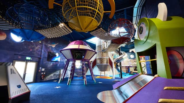 Colorful indoor playground with space-themed structures, slides, and climbing nets under a high blue ceiling.