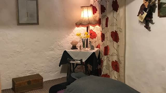 Cozy interior with a bed, floral curtains, a lamp, and a small table with flowers and decor in a rustic setting.