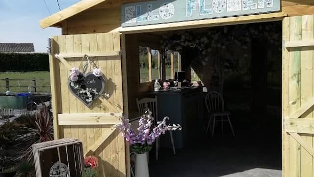 Quaint wooden shed with "Ello Flower" sign, floral decorations, and gravel path in sunny outdoor setting.