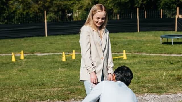 A woman stands with a man kneeling beside a black dog in a grassy area, surrounded by trees and agility cones.