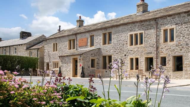 Stone building with large windows, surrounded by flowers and greenery, under a partly cloudy blue sky.