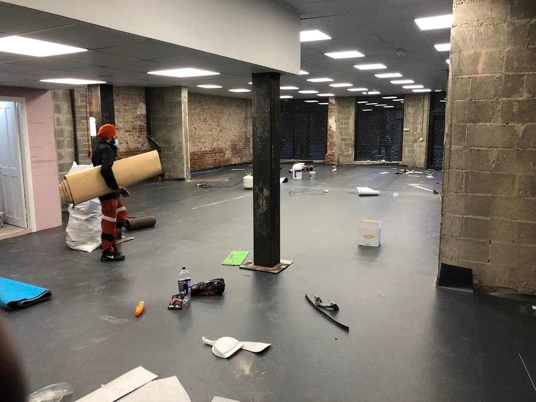 Empty retail space with a worker carrying a roll of carpet, exposed brick walls, and various tools scattered around.