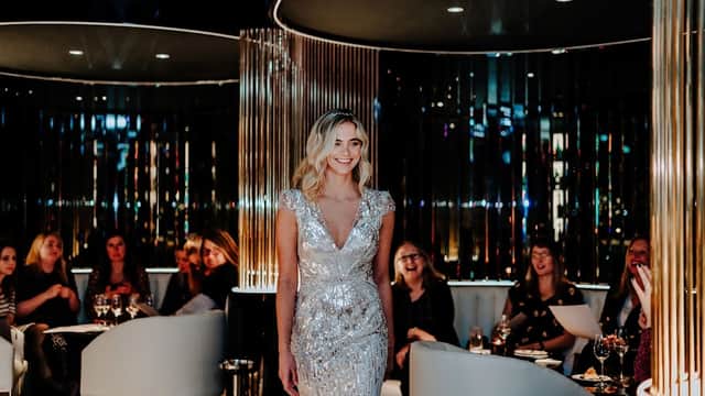 A glamorous woman in a sparkling silver dress walks through a stylish bar, with guests enjoying drinks in the background.