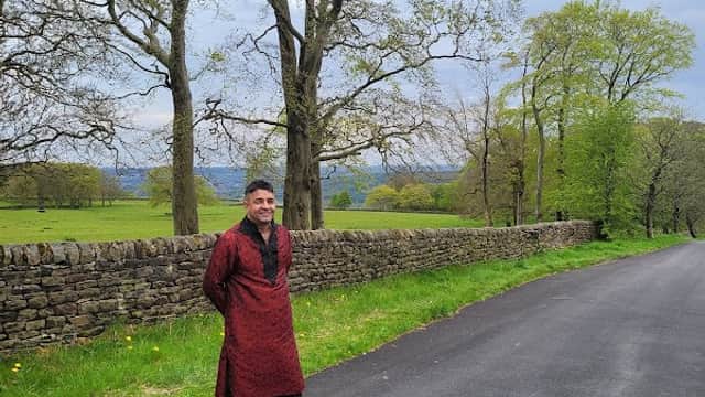 A man in traditional attire stands by a stone wall along a tree-lined road in a lush green landscape.