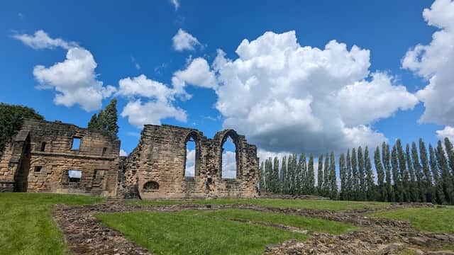 Ruins of Monk Bretton Priory with arched windows, surrounded by green grass and tall trees under a blue sky with clouds.