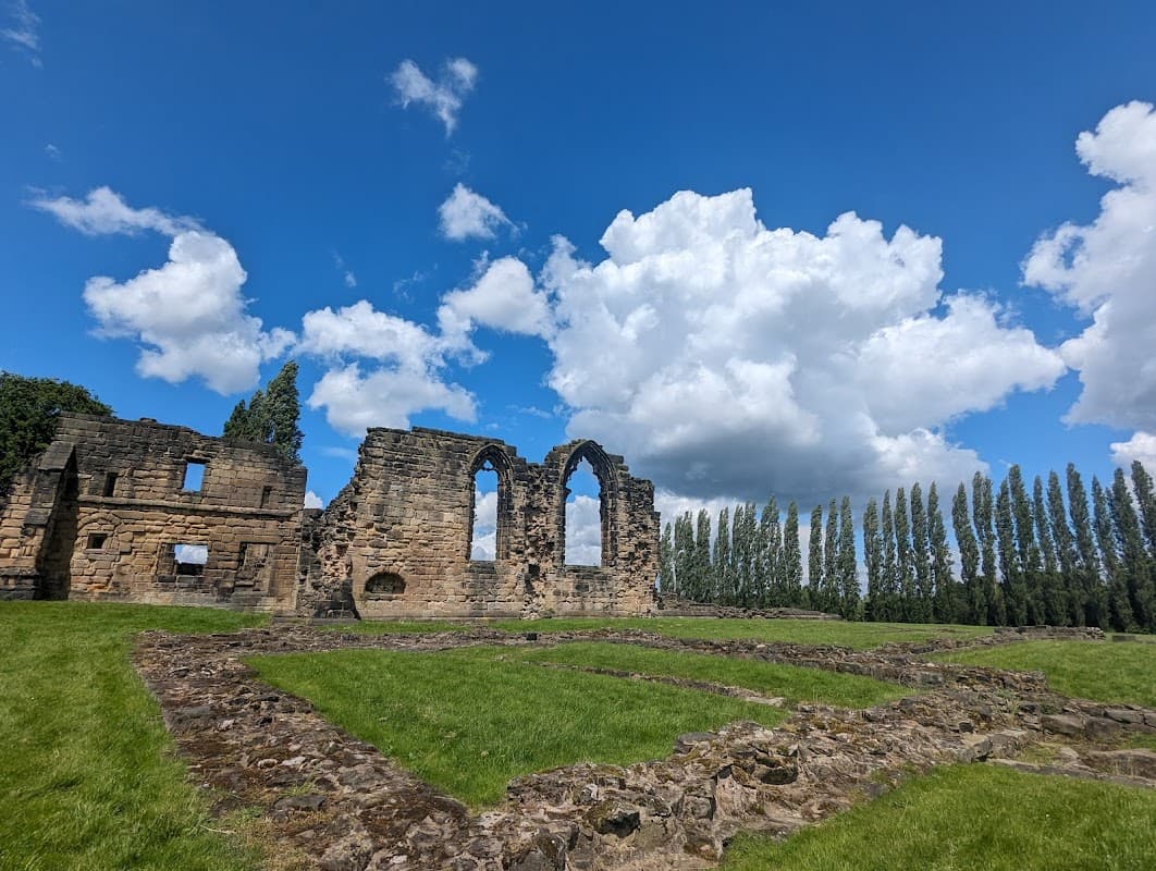 Ruins of Monk Bretton Priory with arched windows, surrounded by green grass and tall trees under a blue sky with clouds.
