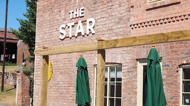 Brick building with "THE STAR" sign, green umbrellas, and wooden tables outside in a sunny setting.