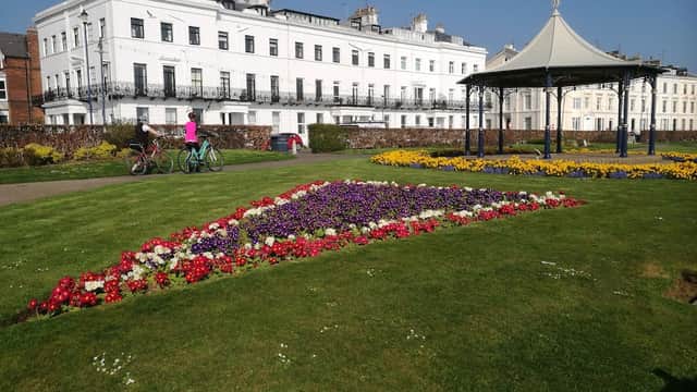 Colorful flower beds in geometric patterns, a gazebo, and people biking near a historic white building.