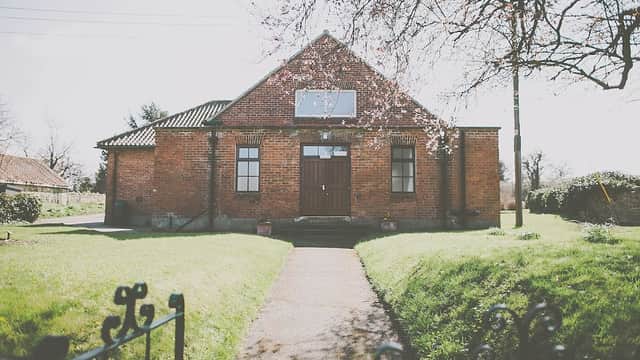 Red brick village hall with a central wooden door, surrounded by green grass and trees under a clear sky.