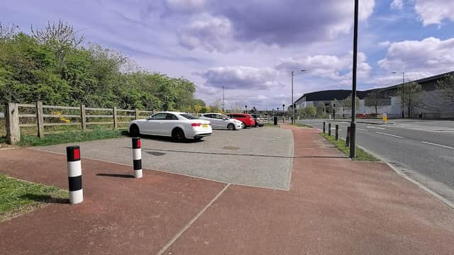 Several parked cars in a gravel car park, surrounded by greenery and a pathway, under a partly cloudy sky.