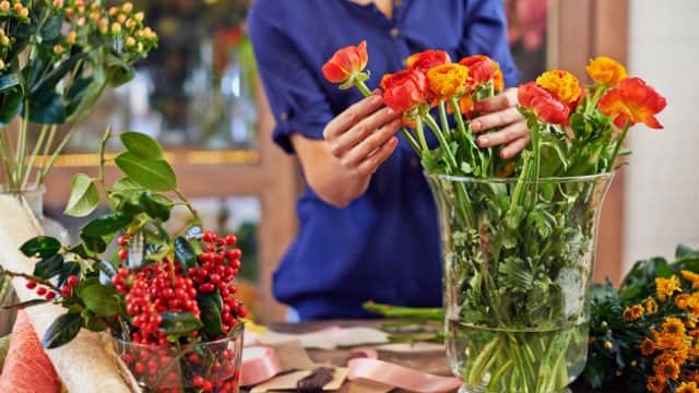 A florist arranging vibrant orange and yellow flowers in a glass vase, surrounded by greenery and decorative materials.