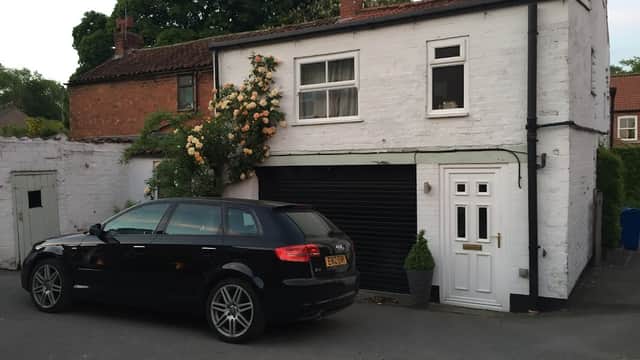 A white two-story building with a black car parked in front, surrounded by greenery and a rose bush.