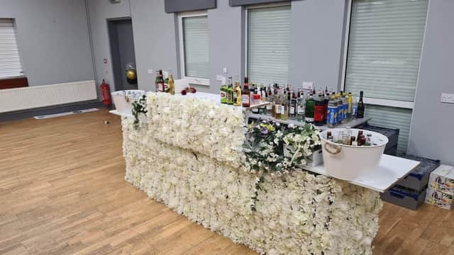 Floral-covered bar with various bottles and drinks, set in a spacious event venue with wooden flooring and large windows.