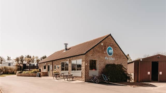 Holiday park building with large windows, outdoor seating, and a bike parked outside in a sunny setting.