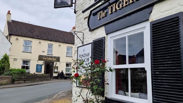 The Tiger Inn sign hangs beside a white building with black shutters, roses in front, and a view of a nearby pub.