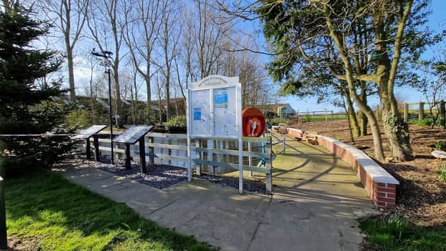 Barmston village pond with informational boards, a lifebuoy, trees, and a path surrounded by green grass.