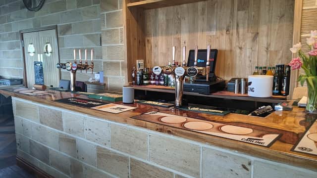 Wooden bar with beer taps, bottles, and glasses; stone wall and floral decorations in a cozy pub setting.