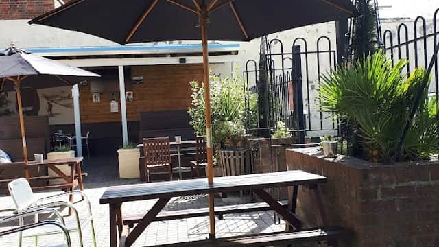 Outdoor seating area with a picnic table under a large umbrella, surrounded by greenery and a fence.