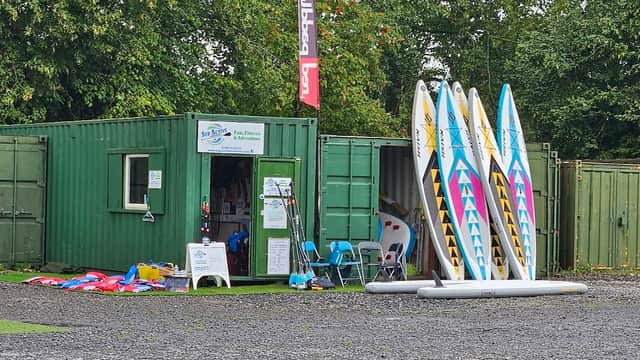 Green shipping container with paddleboards outside, surrounded by trees and equipment for water sports.