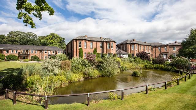 Retirement village with brick buildings, lush greenery, and a serene pond under a partly cloudy sky.