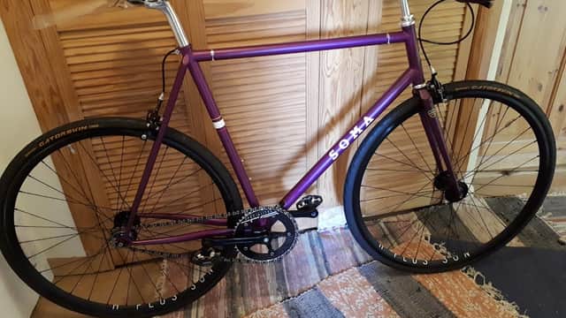Purple fixed-gear bike with black wheels, leaning against a wooden wall, on a patterned rug.