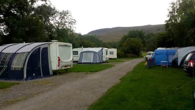Campsite with tents and caravans on a grassy area, surrounded by trees and hills in the background.