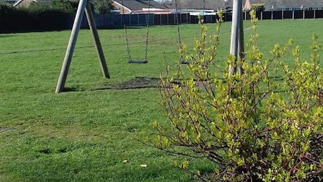 Swing set on a grassy field with a bush in the foreground and houses visible in the background under a clear blue sky.