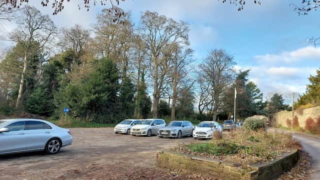 Several parked cars in a gravel lot surrounded by trees and a stone wall under a partly cloudy sky.