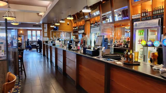 Modern bar area with wooden counters, drinks on display, and patrons seated in a cozy restaurant setting.