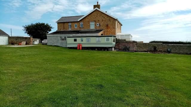 A green caravan on a grassy field beside a large stone house under a blue sky with scattered clouds.