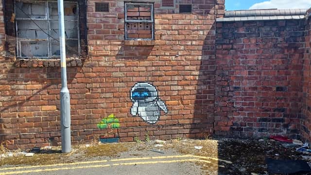 Graffiti of a ghost-like figure on a brick wall, surrounded by litter and overgrown weeds in a car park.