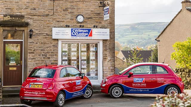 Two red cars parked outside a stone building with a sign for "Wilman & Lodge" and property listings in the windows.