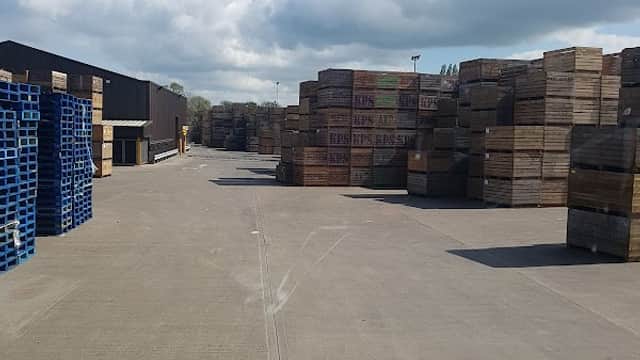Stacks of wooden pallets and crates lined along a concrete pathway under a partly cloudy sky at a produce facility.