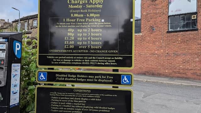 Sign for Queens Road car park with parking charges and conditions, surrounded by buildings and a road in Halifax, Yorkshire.
