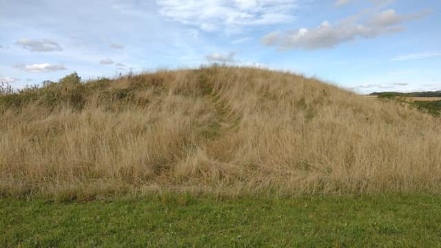 A grassy mound surrounded by tall grass under a blue sky with scattered clouds in Duggleby, Yorkshire.