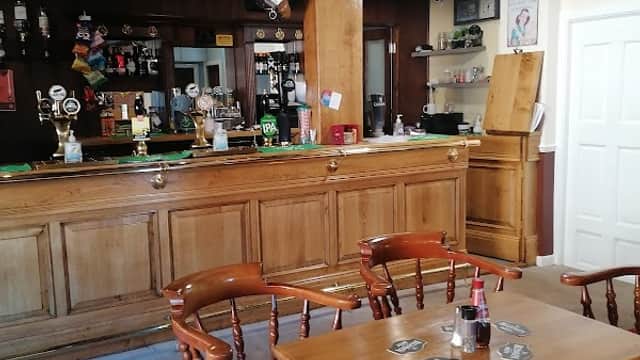 Wooden bar with taps, stools, and tables; rustic decor featuring dark beams and framed pictures in a cozy pub setting.
