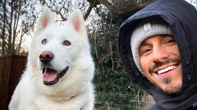 A man in a black jacket smiles beside a fluffy white dog with blue eyes in a park setting.