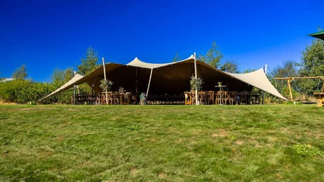 Large tent set up on green grass, surrounded by trees, with wooden tables and chairs arranged inside. Clear blue sky.