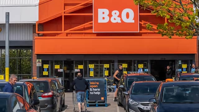 B&Q store entrance with customers, parked cars, and a sign stating "We're open & here to help."