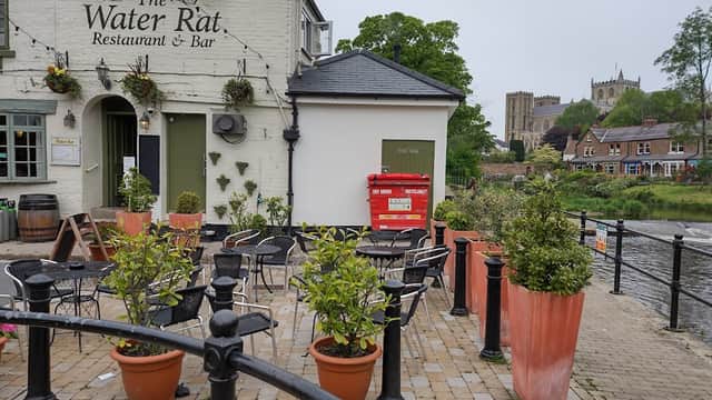 The Water Rat bar with outdoor seating, potted plants, and a view of a church in the background by the river.